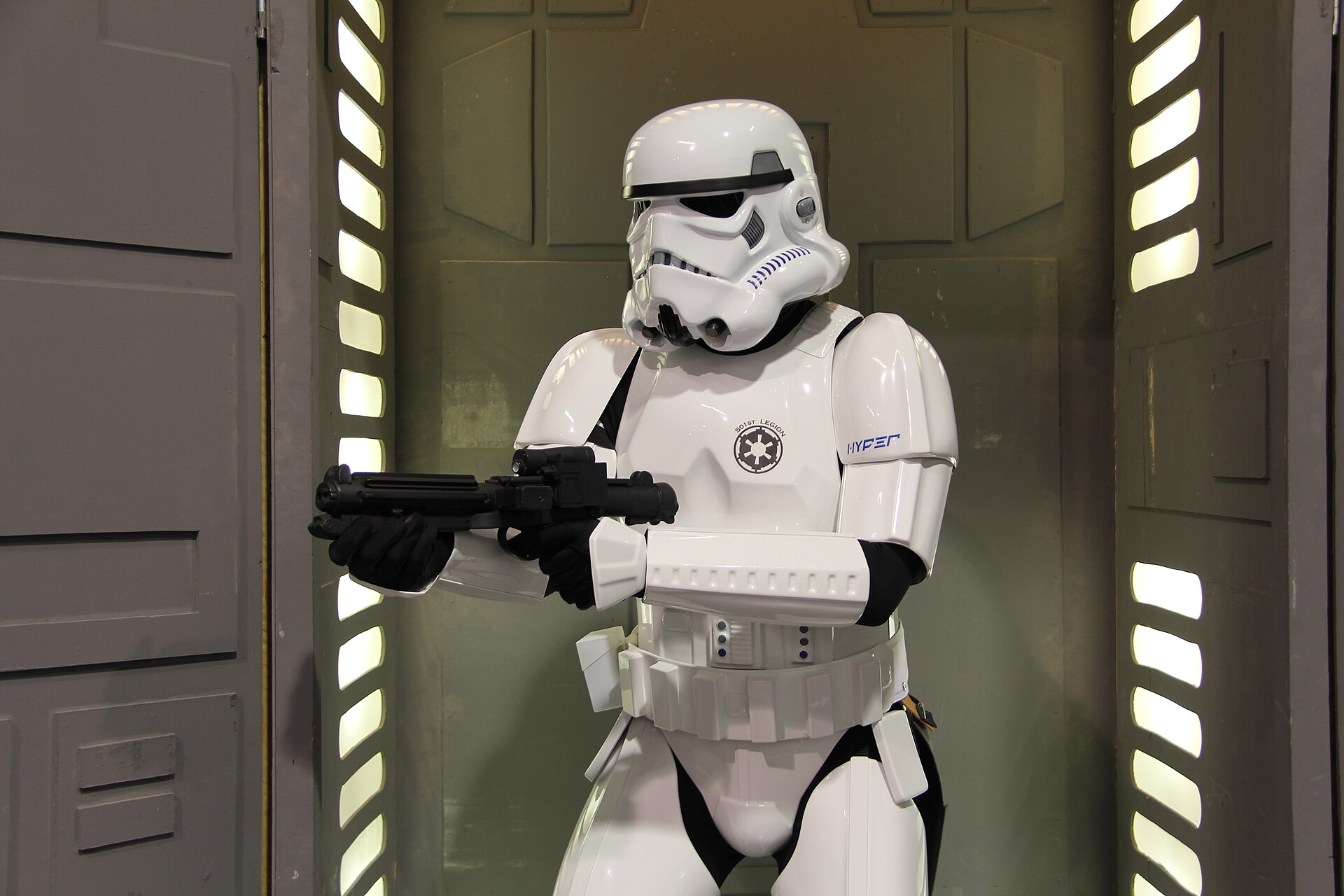 A costumed stormtrooper aiming a blaster inside a lit, spaceship-style corridor set at Star Wars Celebration