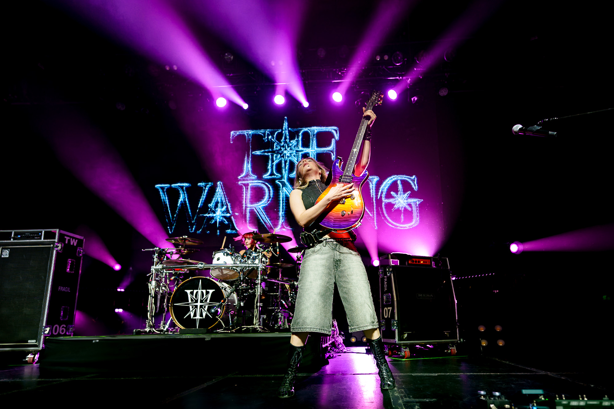The Warning guitarist raising her guitar under purple stage lights at The Fonda Theatre in Los Angeles.