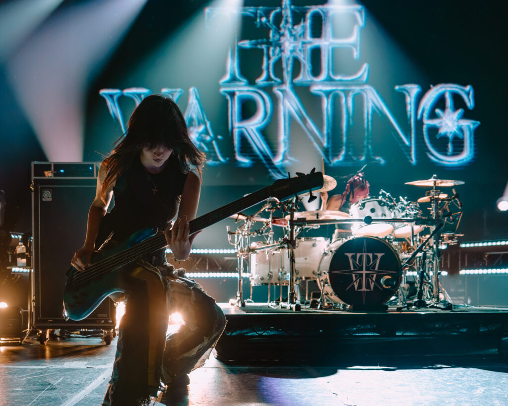 The Warning bassist Ale Villarreal performing at The Fonda Theatre in Los Angeles, with blue neon LIVE letters behind her and the drum kit in the background.