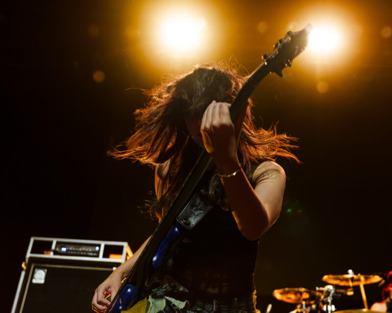 The Warning guitarist playing a blue electric guitar under bright stage lights at The Fonda Theatre in Los Angeles.