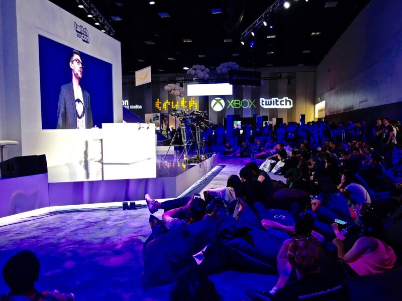 A speaker at a podium on a purple-lit TwitchCon stage, shown on a large screen, with an audience seated on the floor of the San Diego Convention Center