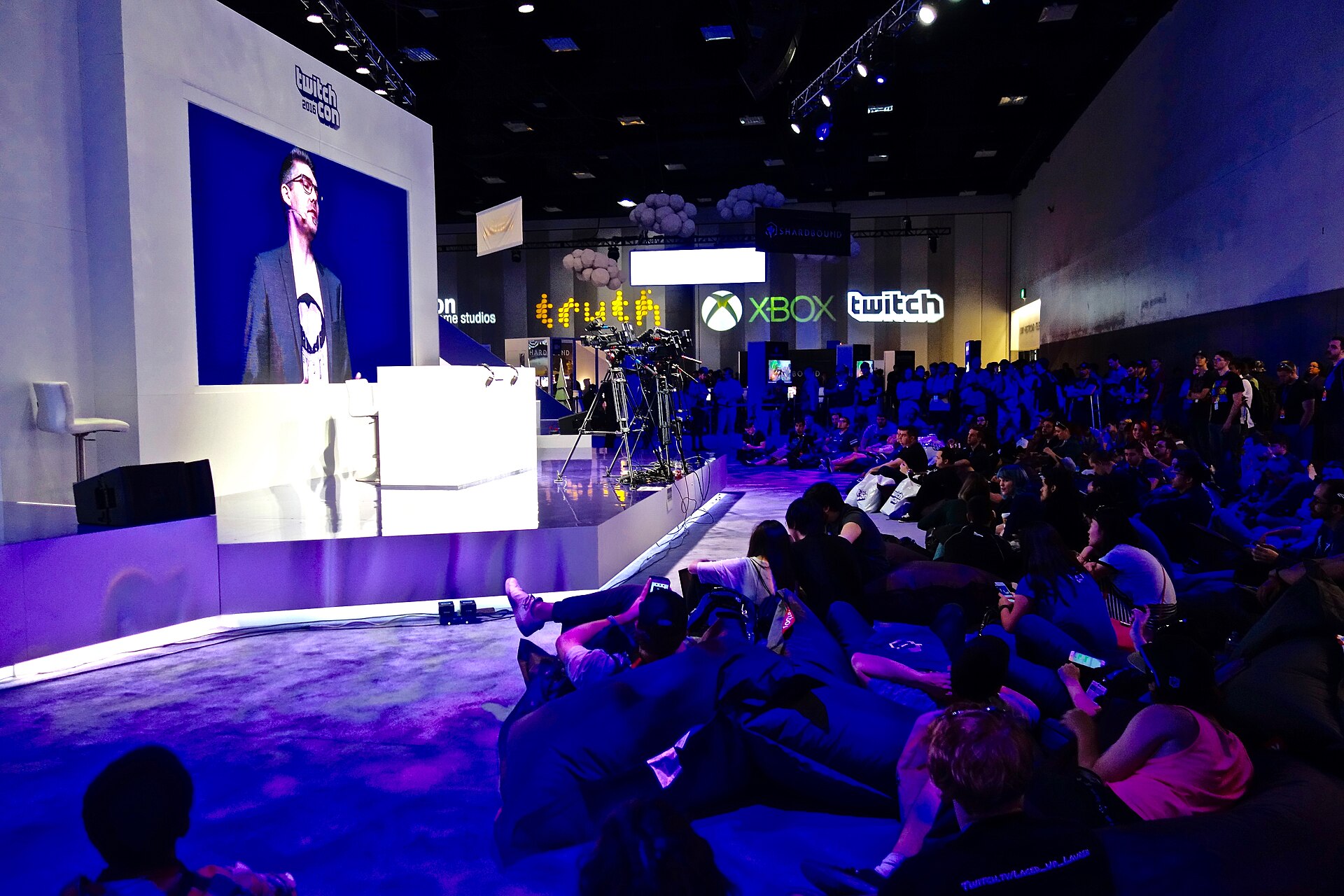 A speaker at a podium on a purple-lit TwitchCon stage, shown on a large screen, with an audience seated on the floor of the San Diego Convention Center