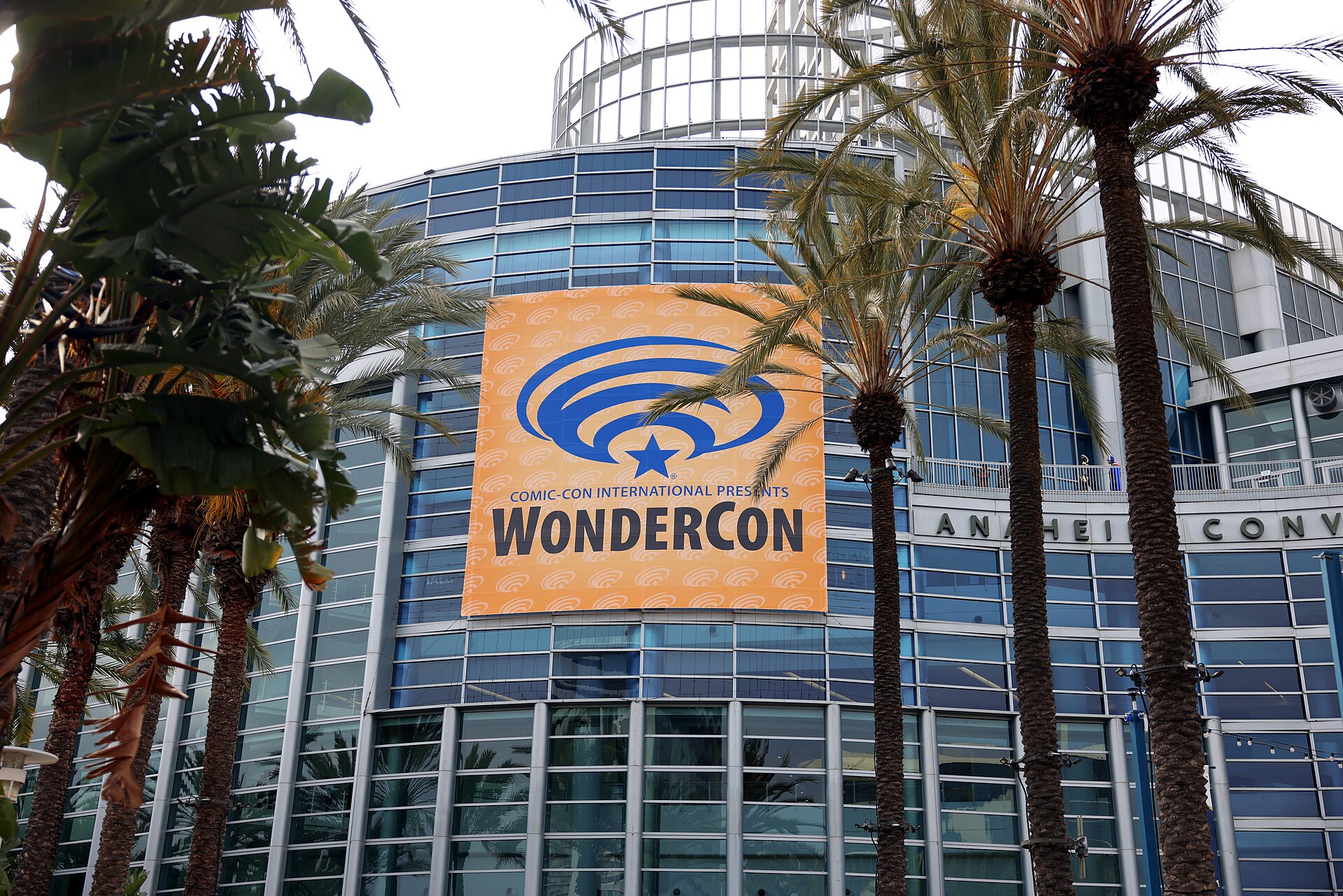 A large WonderCon banner hanging on the curved glass front of the Anaheim Convention Center, framed by palm trees