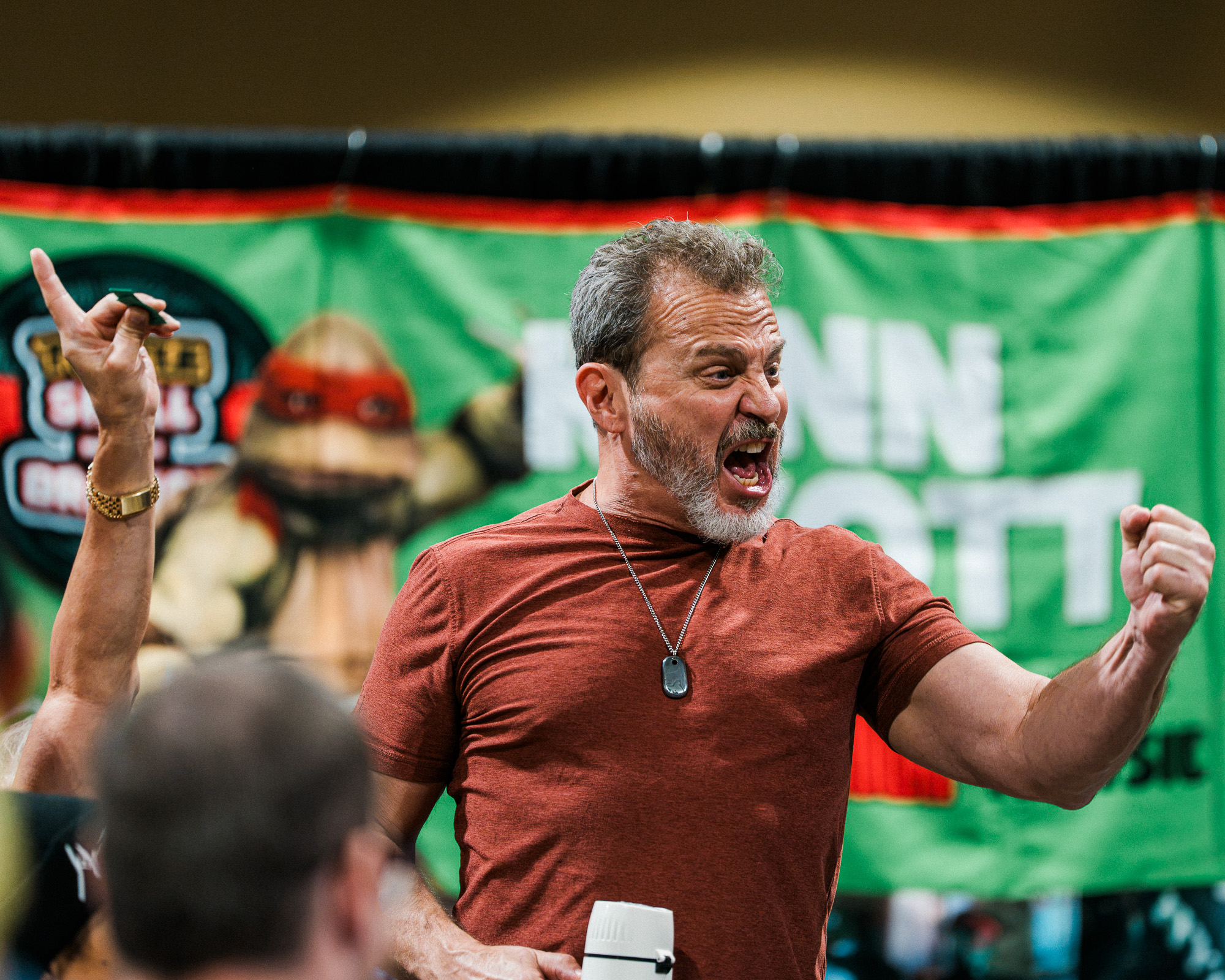 Long Beach Comic Con 2026: man with gray beard in a red shirt shouting with arms raised at a public event, green banner in background.