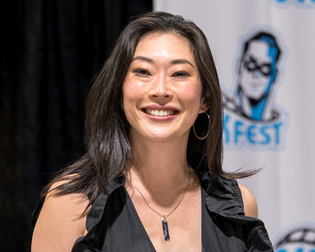 Zharmaine-Boatman-Long-Beach-Comic-Con-the8244journalpress-46885 – The 824 Journal Smiling woman with long dark hair wearing a black sleeveless top and a pendant necklace at an event, backdrop with a logo.