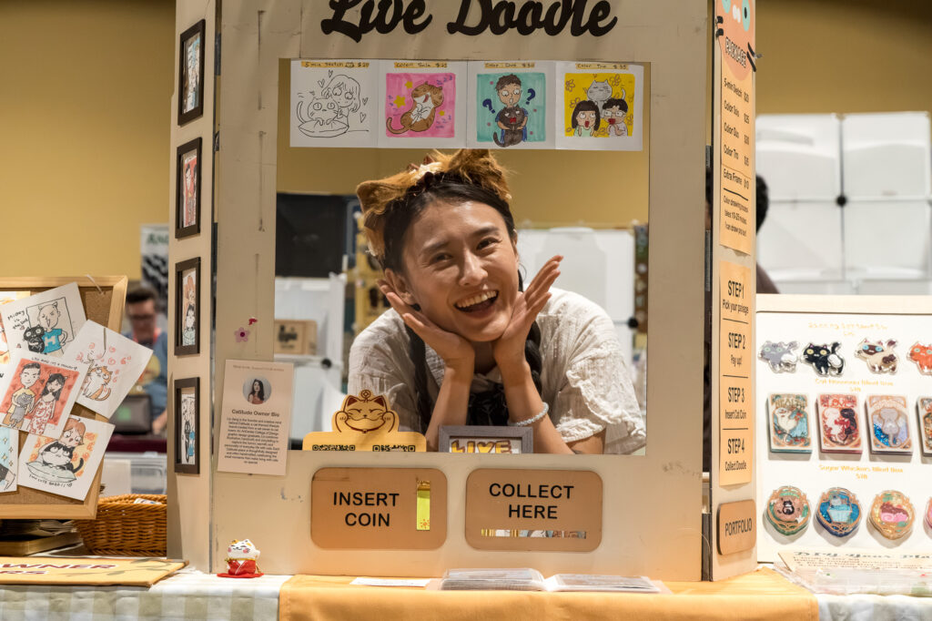 Zharmaine-Boatman-Long-Beach-Comic-Con-the8244journalpress-46901 – The 824 Journal Smiling woman inside a doodle booth labeled 'Live Doodle' with cartoon drawings on the walls and a coin slot reading 'Insert Coin/Collect Here' in front of her.