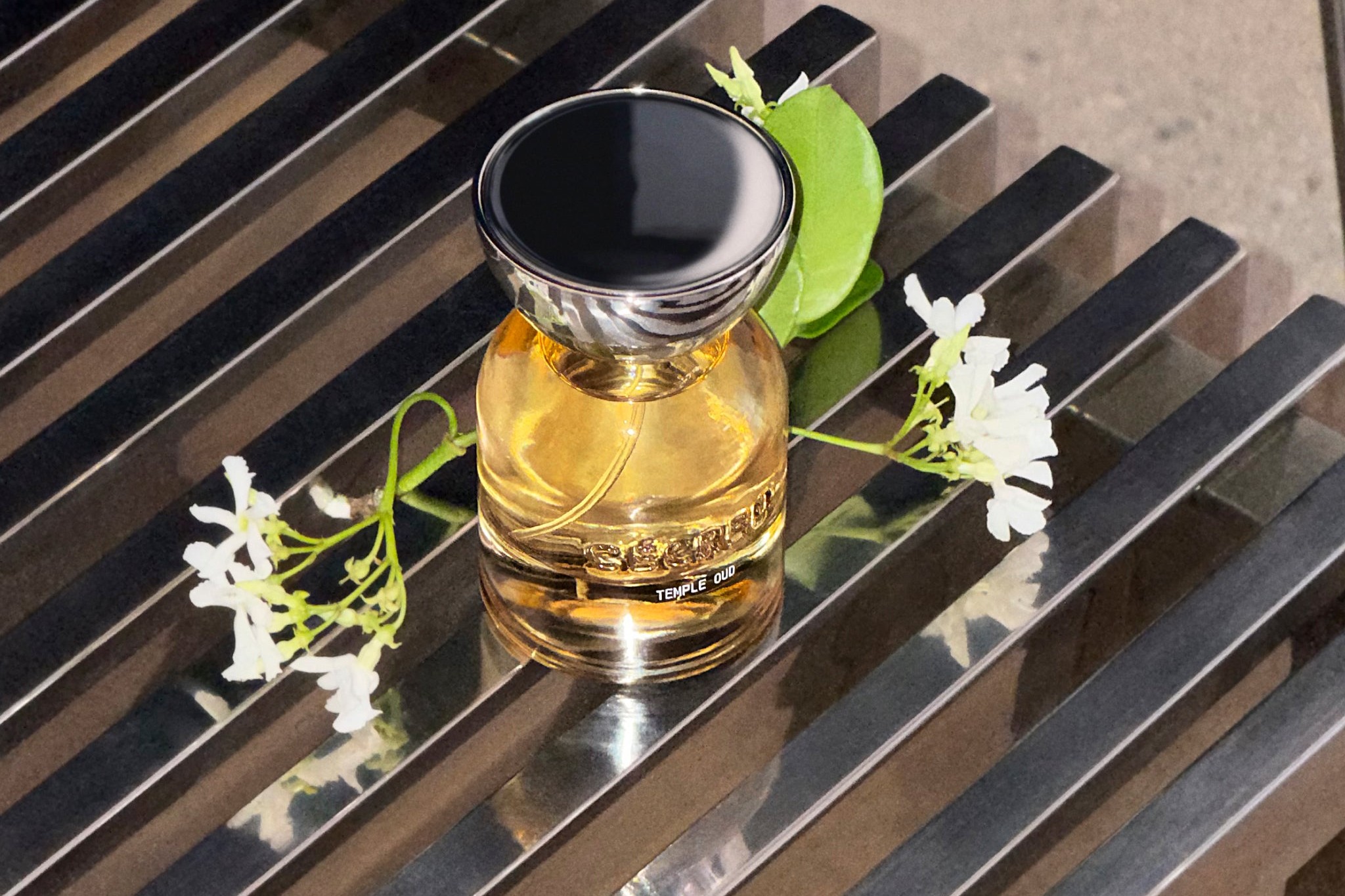 Cécred Temple Oud Hair Perfume in an amber glass bottle with a chrome cap, styled with white jasmine flowers and a green leaf