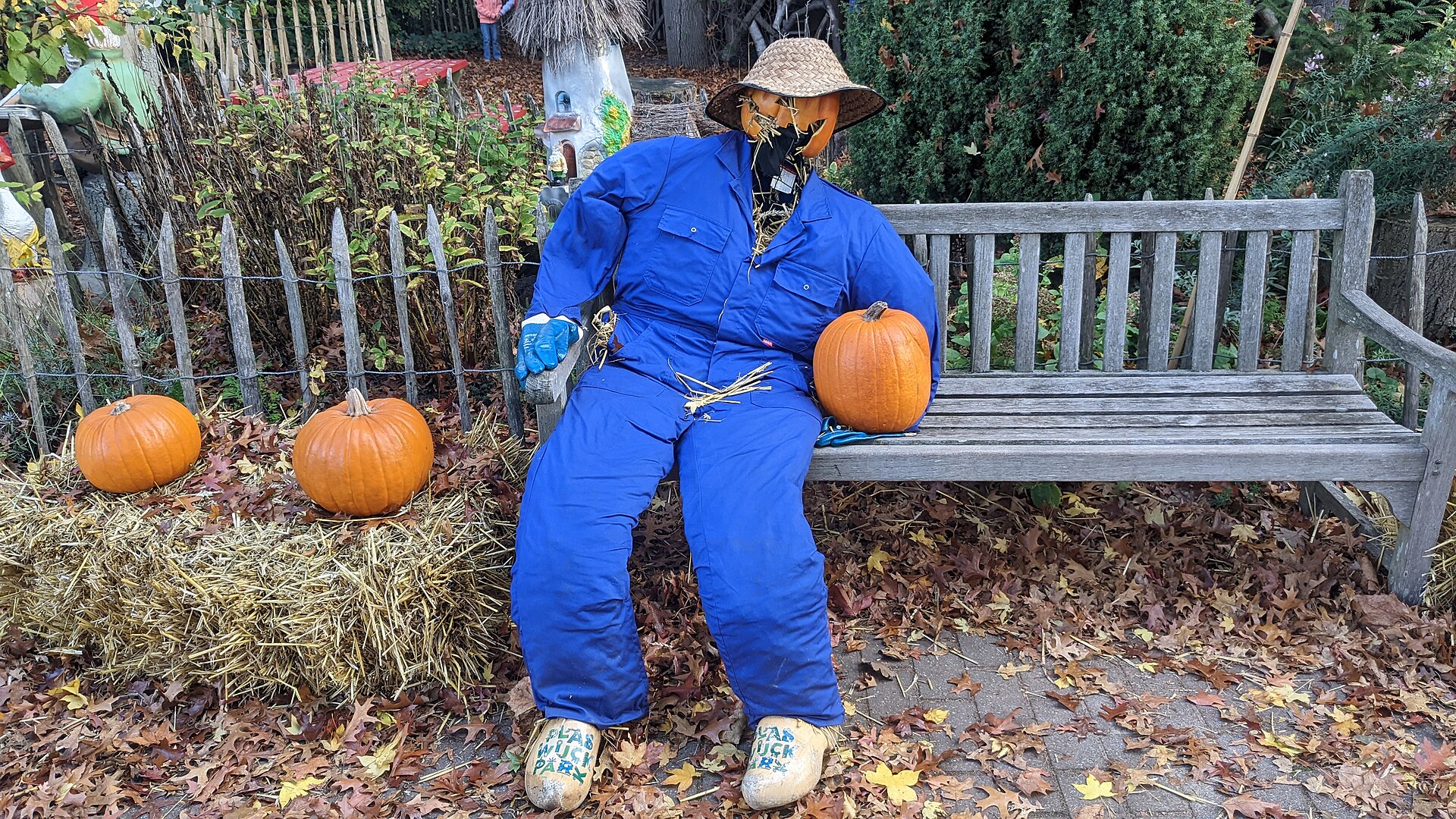 A scarecrow in blue overalls and a straw hat sits on a park bench beside orange pumpkins and hay