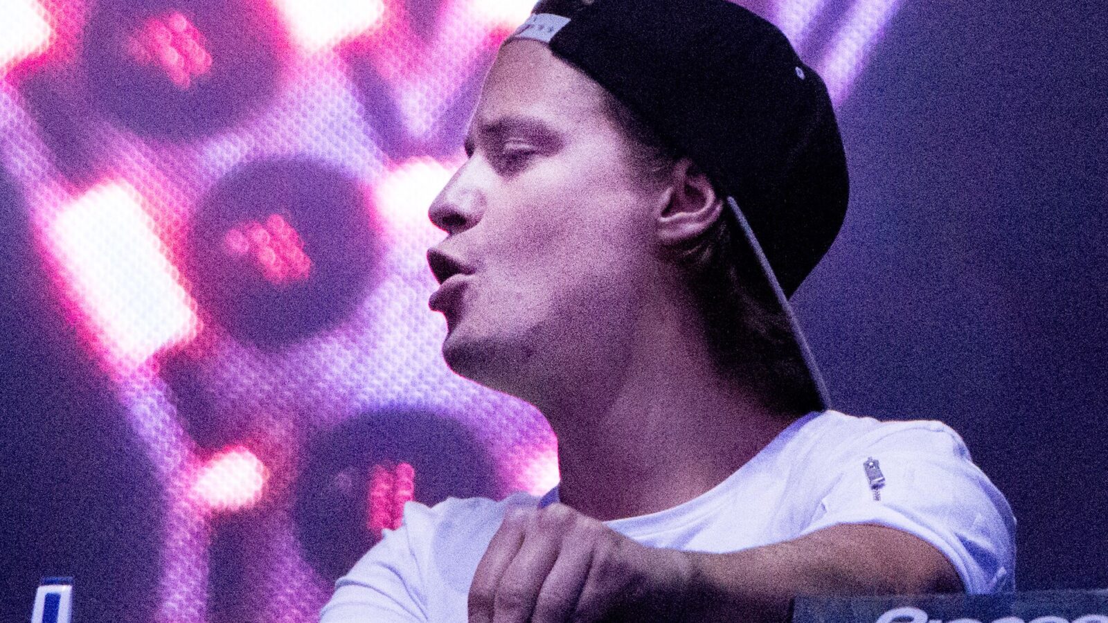 Kygo behind the decks in a white cap during a DJ set, with pink stage lights behind him
