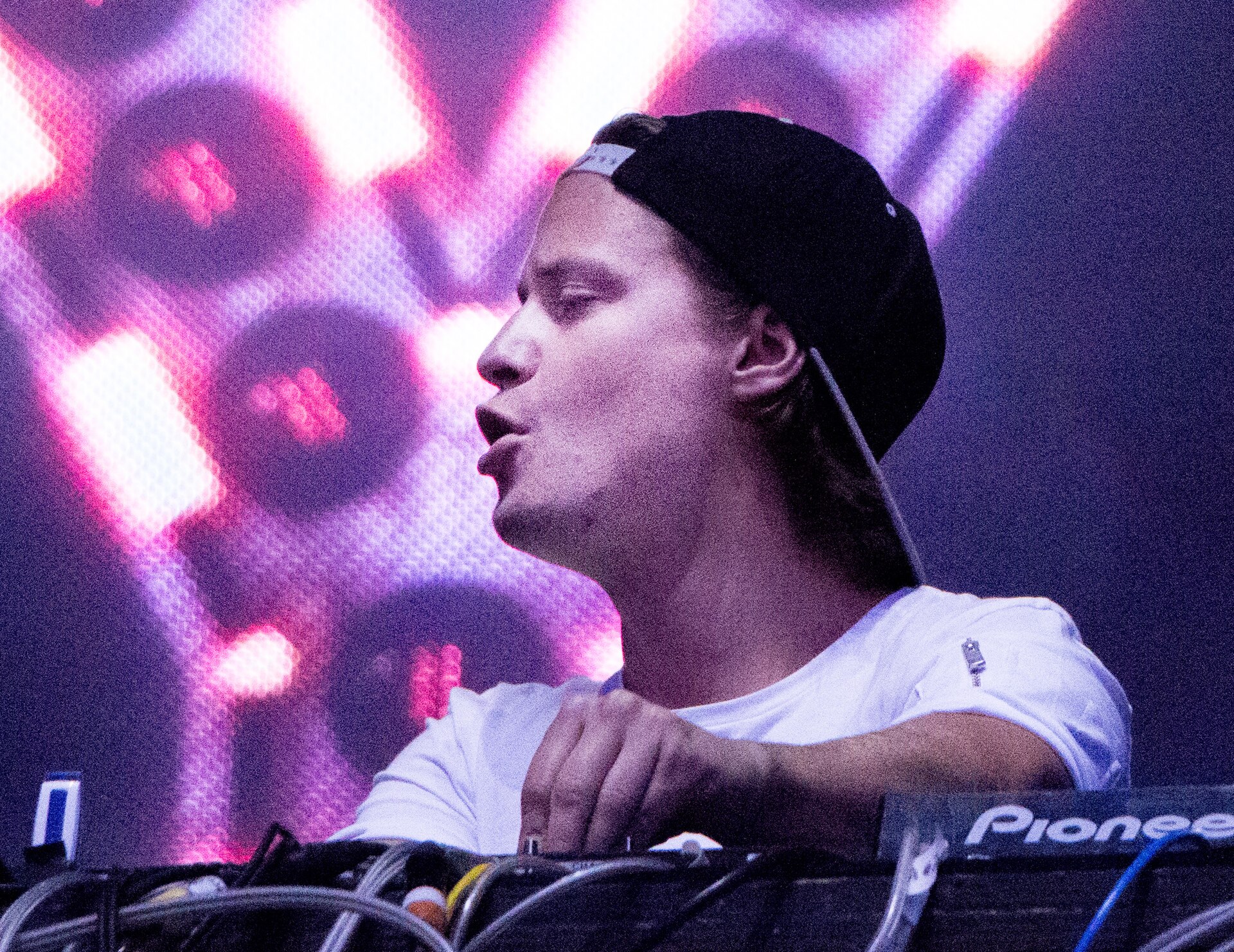 Kygo behind the decks in a white cap during a DJ set, with pink stage lights behind him